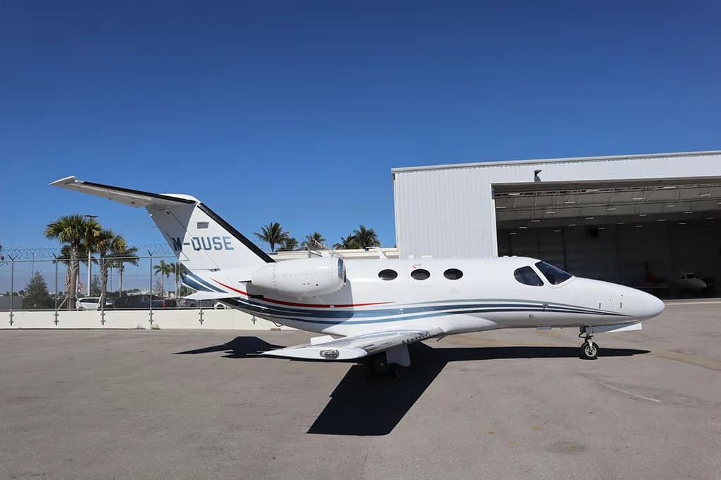 A white business jet with blue and red stripes, seen from the side, parked on an apron with palm trees and a hangar in the background.
