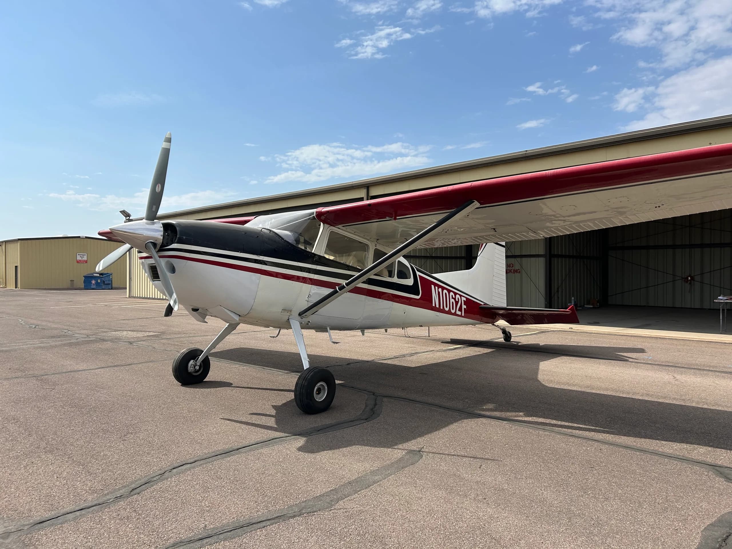 A white, black, and red single-engine, high-wing propeller aircraft (N1062F) is parked on an airfield next to a hangar under a partly cloudy sky.