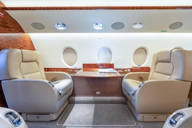 Two light-colored leather seats facing each other across a polished wood table in the mid-cabin of a private jet.