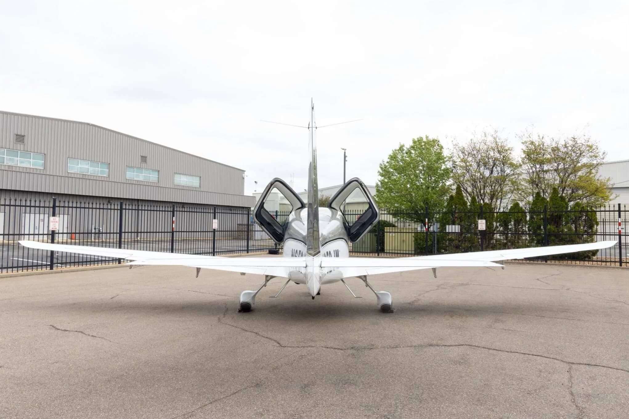A rear view of a silver and white Cirrus SR22 aircraft with both gull-wing doors open, parked on an tarmac.
