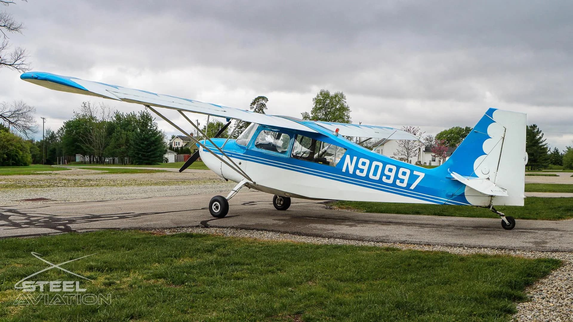 Side view of a white and blue Bellanca 7GCBC Citabria aircraft with registration N90997, parked on an asphalt ramp with trees in the background.