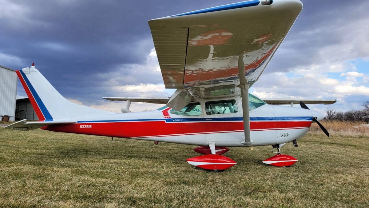 A side view of the Cessna Skylane N3291S with red, white, and blue paint scheme on a grassy field.