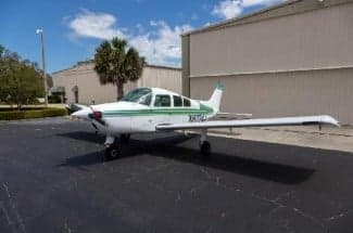 A white and green Mooney M20C aircraft is parked on the tarmac in front of a hangar.