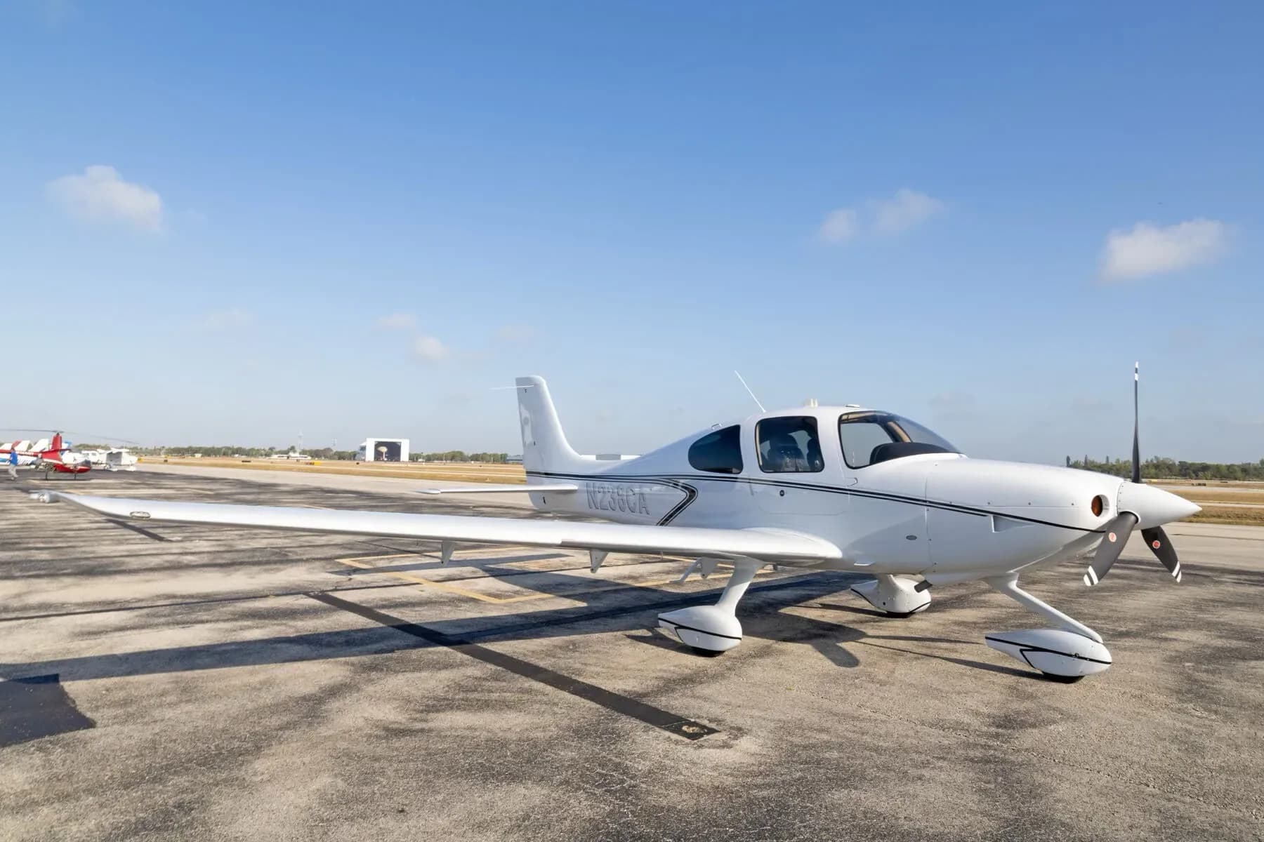 A white Cirrus SR22 aircraft with black stripes is parked on an airport tarmac under a blue sky.