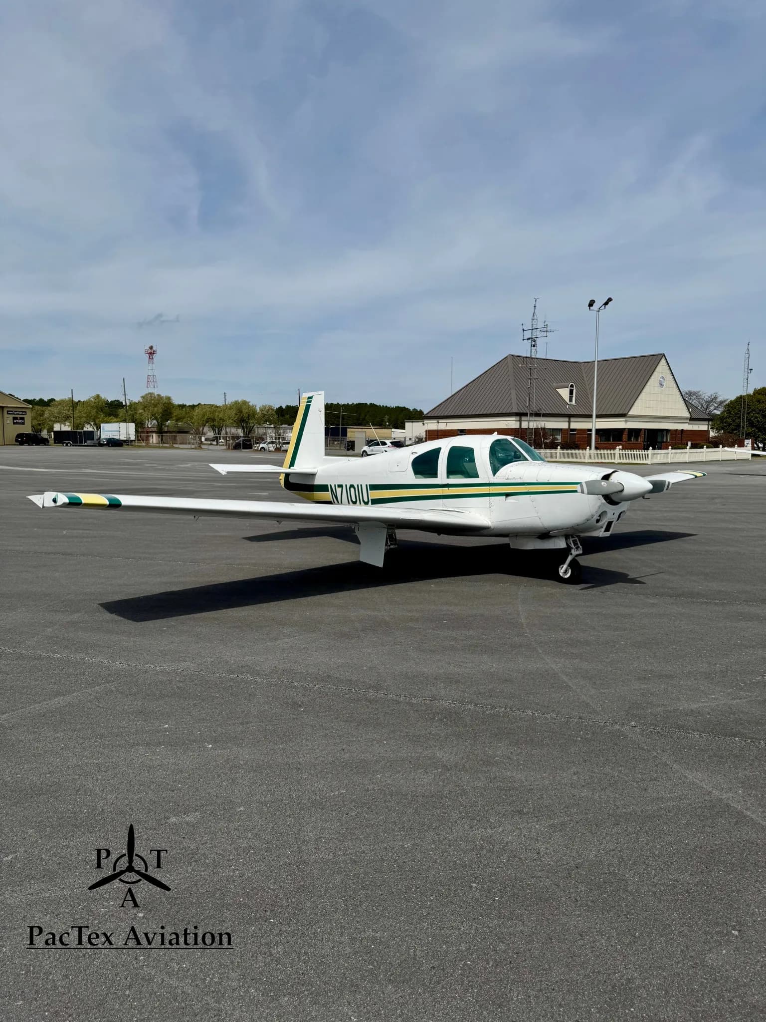 A white Mooney M20C aircraft with green and yellow stripes, parked on an airport tarmac with a building in the distance.