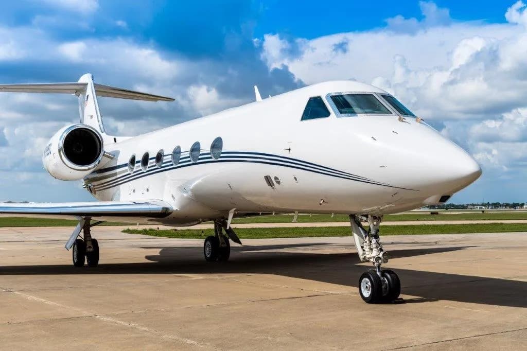 A white Gulfstream business jet with blue stripes is parked on an airport tarmac under a partly cloudy sky.