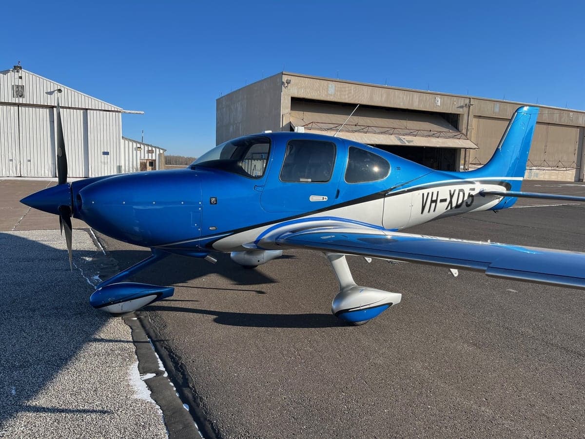 A blue and white Cirrus SR22 aircraft parked on the tarmac in front of a hangar.