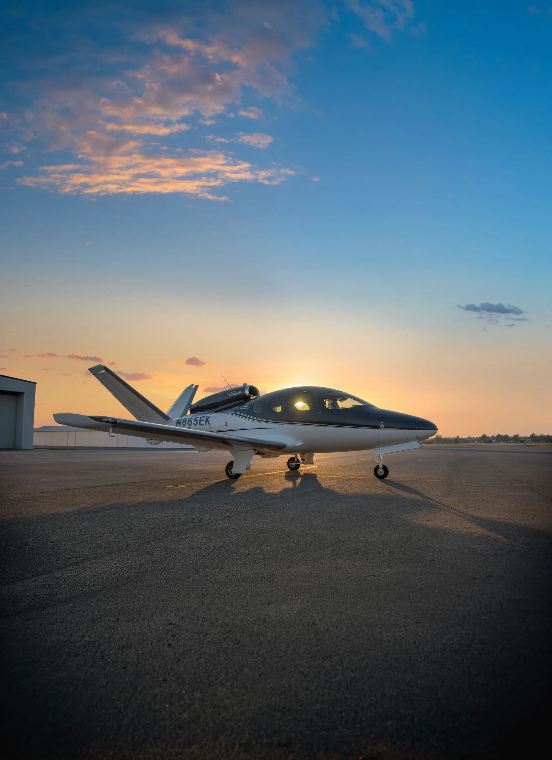 A Cirrus Vision Jet (N865EK) with a dark grey and white paint scheme is parked on an airfield at sunset.
