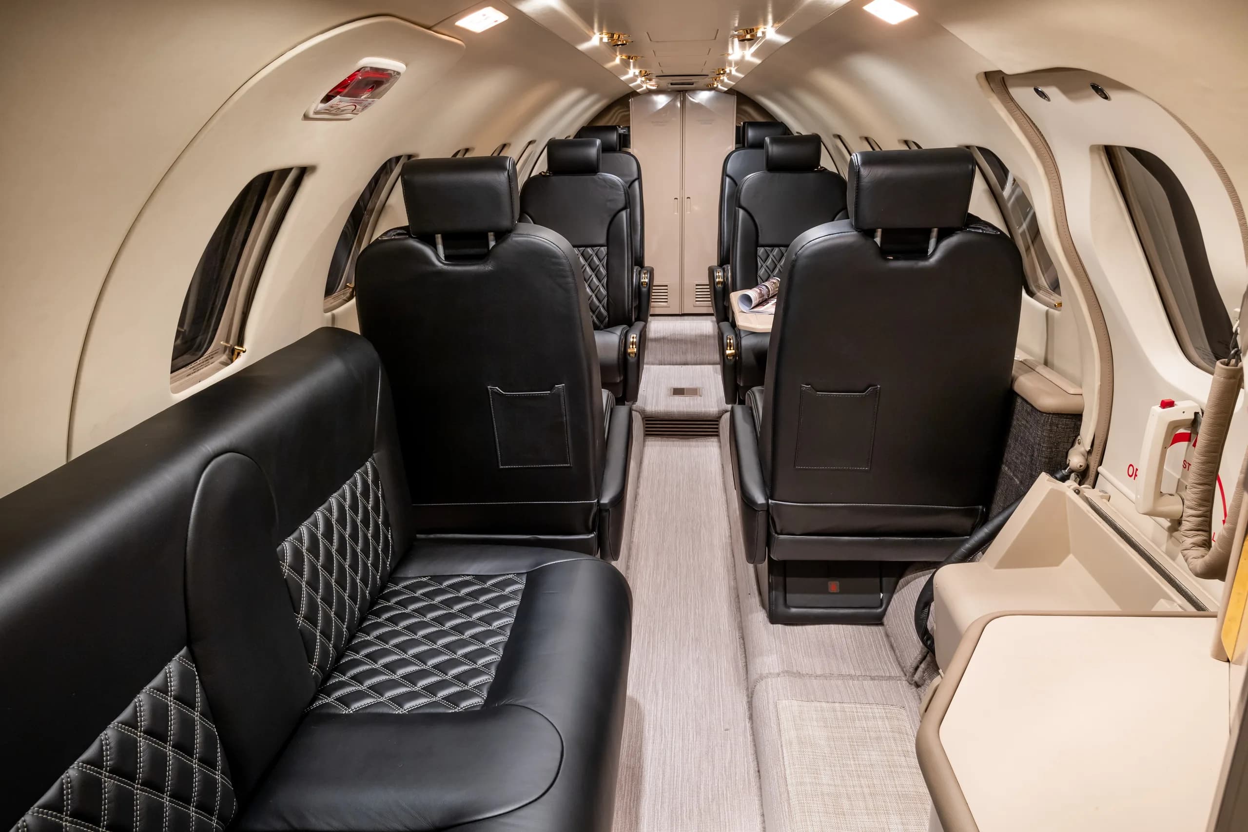 The interior cabin of a business jet, looking towards the rear, with black quilted leather seats and light-colored walls.