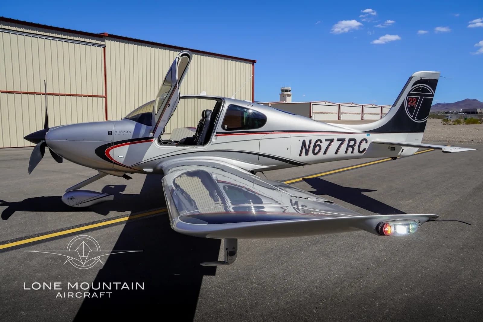A silver Cirrus SR22 aircraft with red and black accents, registration N677RC, parked on an airfield with its left door open, showing the tail with a "22 G5" logo.