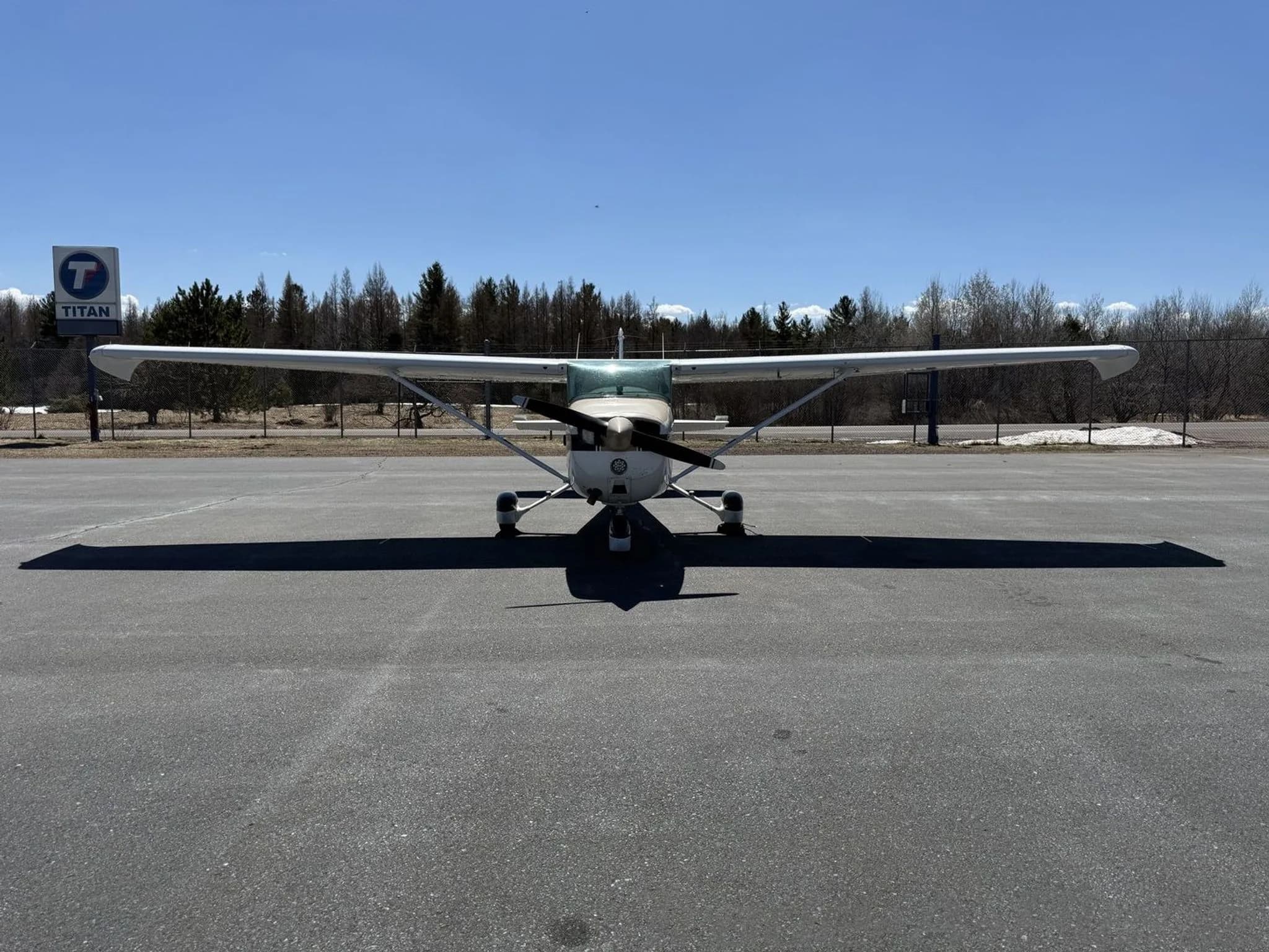 A head-on view of a white and tan Cessna 172 aircraft with blue stripes parked on an asphalt apron.