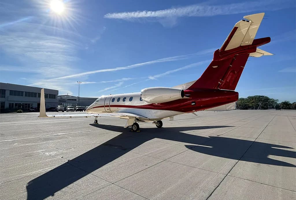 A rear view of a white and red Embraer Phenom 300 private jet on a tarmac with sun glare.
