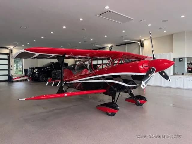 A red and black biplane with a checkered pattern, viewed from the front-right, parked in a garage.