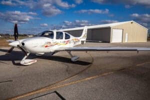 A Cirrus SR22 aircraft with a custom paint scheme is parked on the tarmac in front of a hangar.