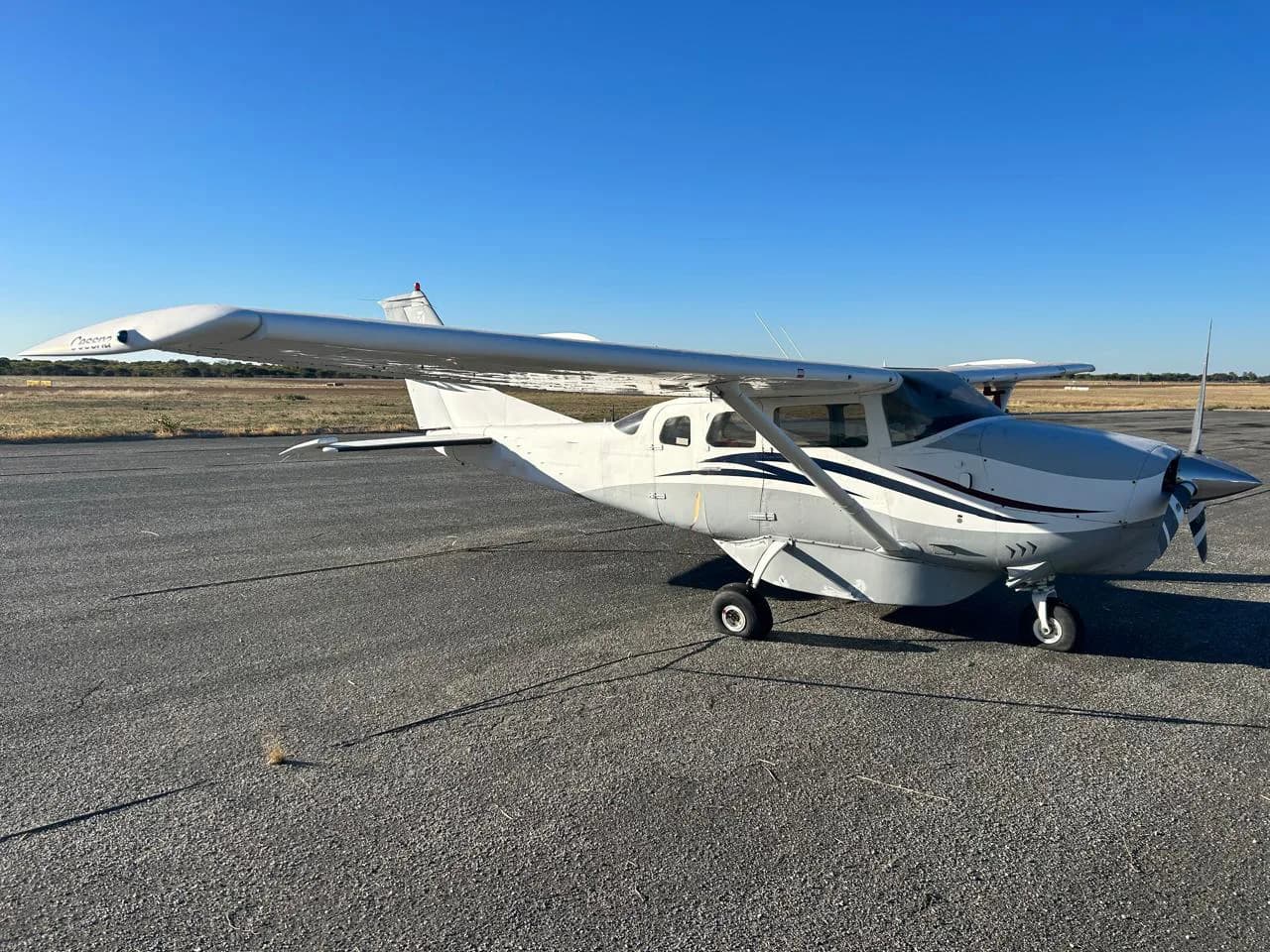 A white and grey single-engine high-wing aircraft parked on a tarmac under a clear blue sky.