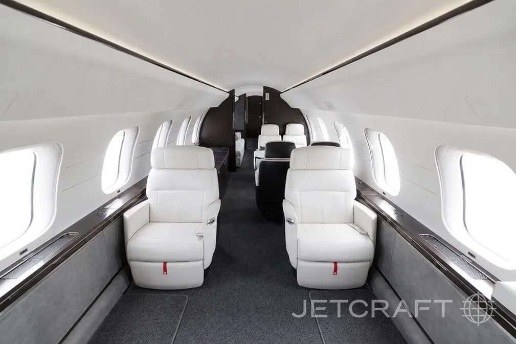 A view down the aisle of a luxurious aircraft cabin with white leather seats and large windows.