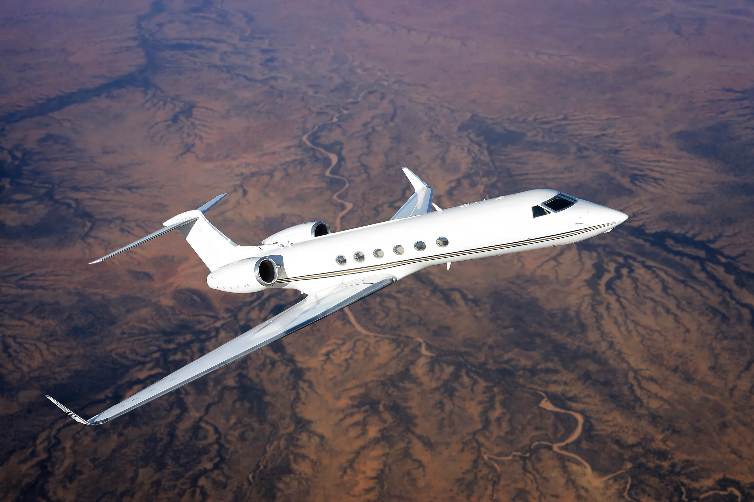 A white business jet with a gold stripe flying over a desert landscape.
