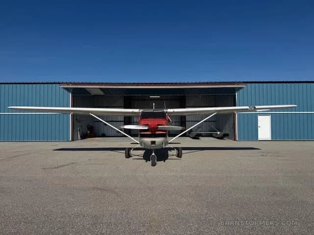 A red and white Cessna 172, registration N2835L, viewed head-on, parked on an asphalt apron in front of a blue hangar.