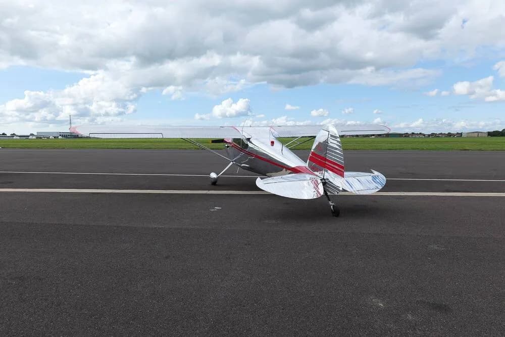 A rear view of a silver Cessna 170B with red trim, parked on an airfield under a partly cloudy sky.