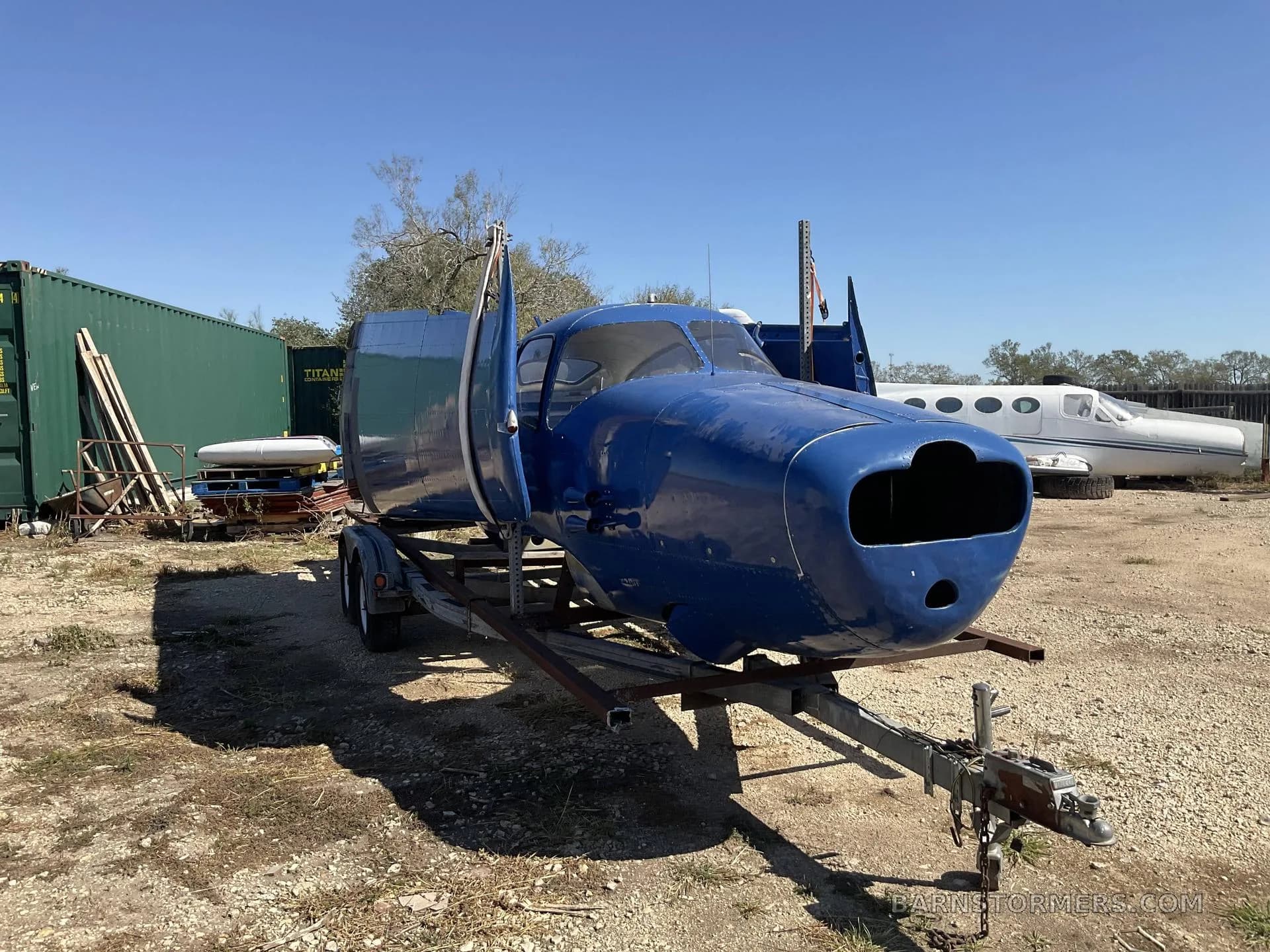 A blue aircraft fuselage on a trailer, with another white aircraft fuselage visible in the background, in a sunny gravel lot.