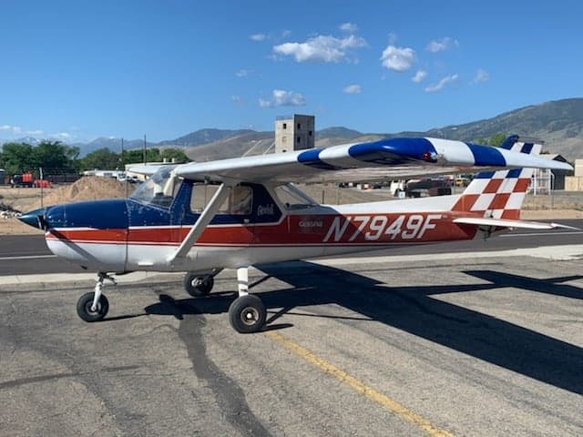 A Cessna 150/152 aircraft with a red, white, and blue paint scheme, including a checkered tail, parked on an asphalt ramp with mountains in the background.