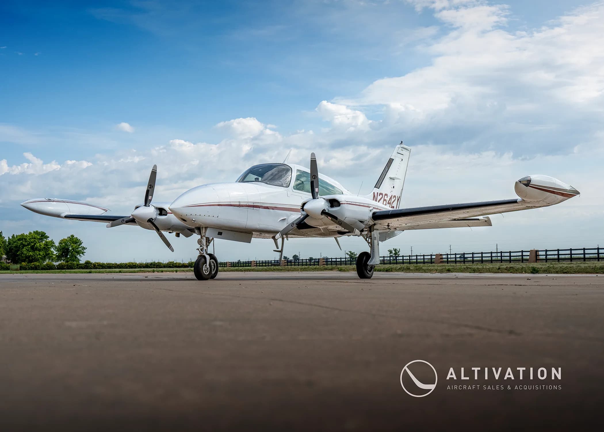 A low-angle view of a white twin-engine propeller aircraft with maroon stripes, tail number N2642Y, parked on an airfield under a partly cloudy sky.