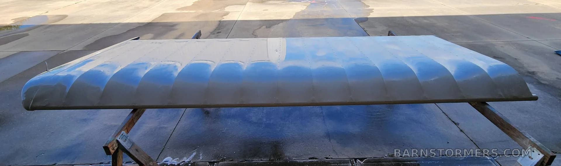 A light grey aircraft wing with a ribbed surface, resting on sawhorses on a wet concrete pad.