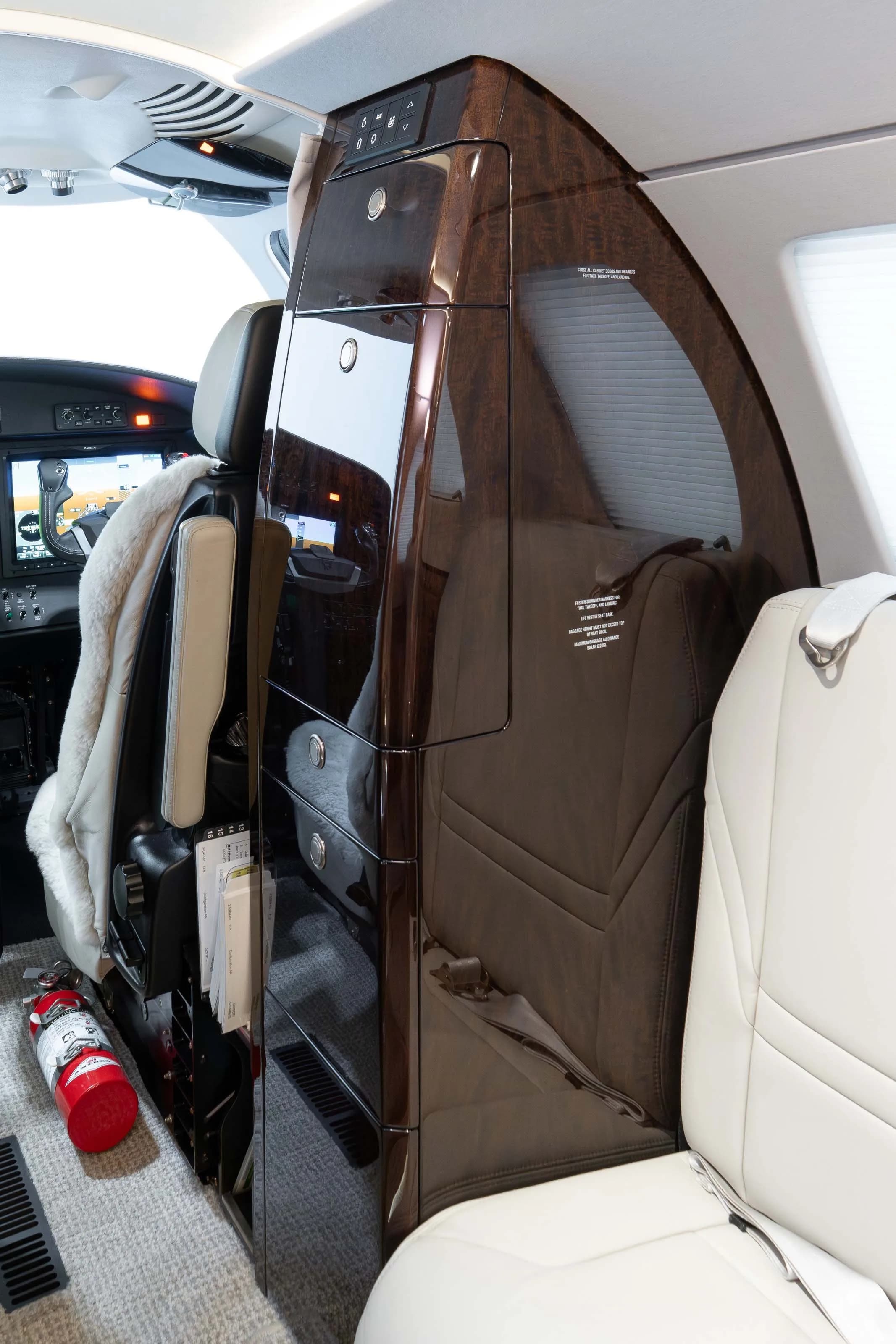 A close-up view of the luxurious dark wood cabinetry and a light grey seat in the business jet's cabin, with a fire extinguisher visible.
