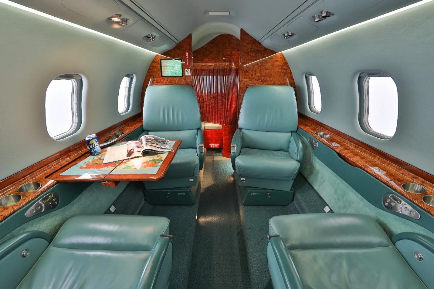 The interior cabin of a private jet with teal leather seats, wood paneling, and a table with a magazine and drink.