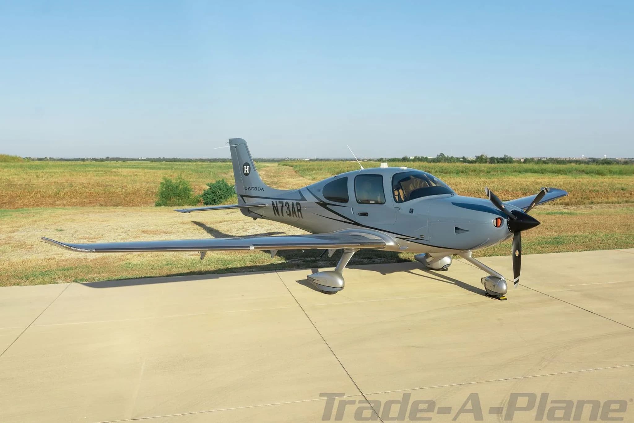 A side view of a grey Cirrus SR22 aircraft with registration N73AR parked on a tarmac next to a grassy field.