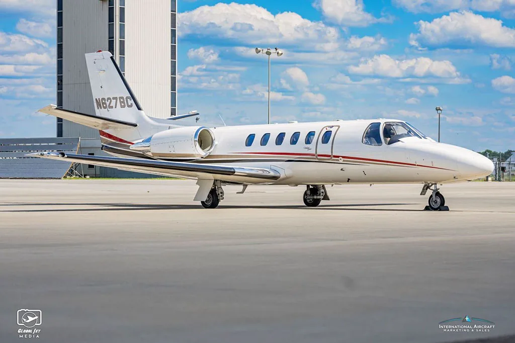 A side view of a white Cessna Citation Bravo business jet with red and black stripes parked on the tarmac.