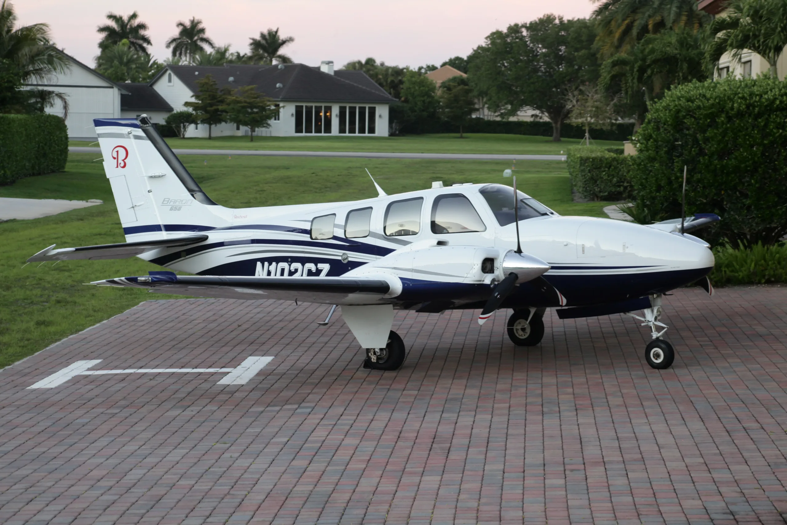 A white and blue Beechcraft Baron N102CZ parked on a paved driveway with houses in the background.