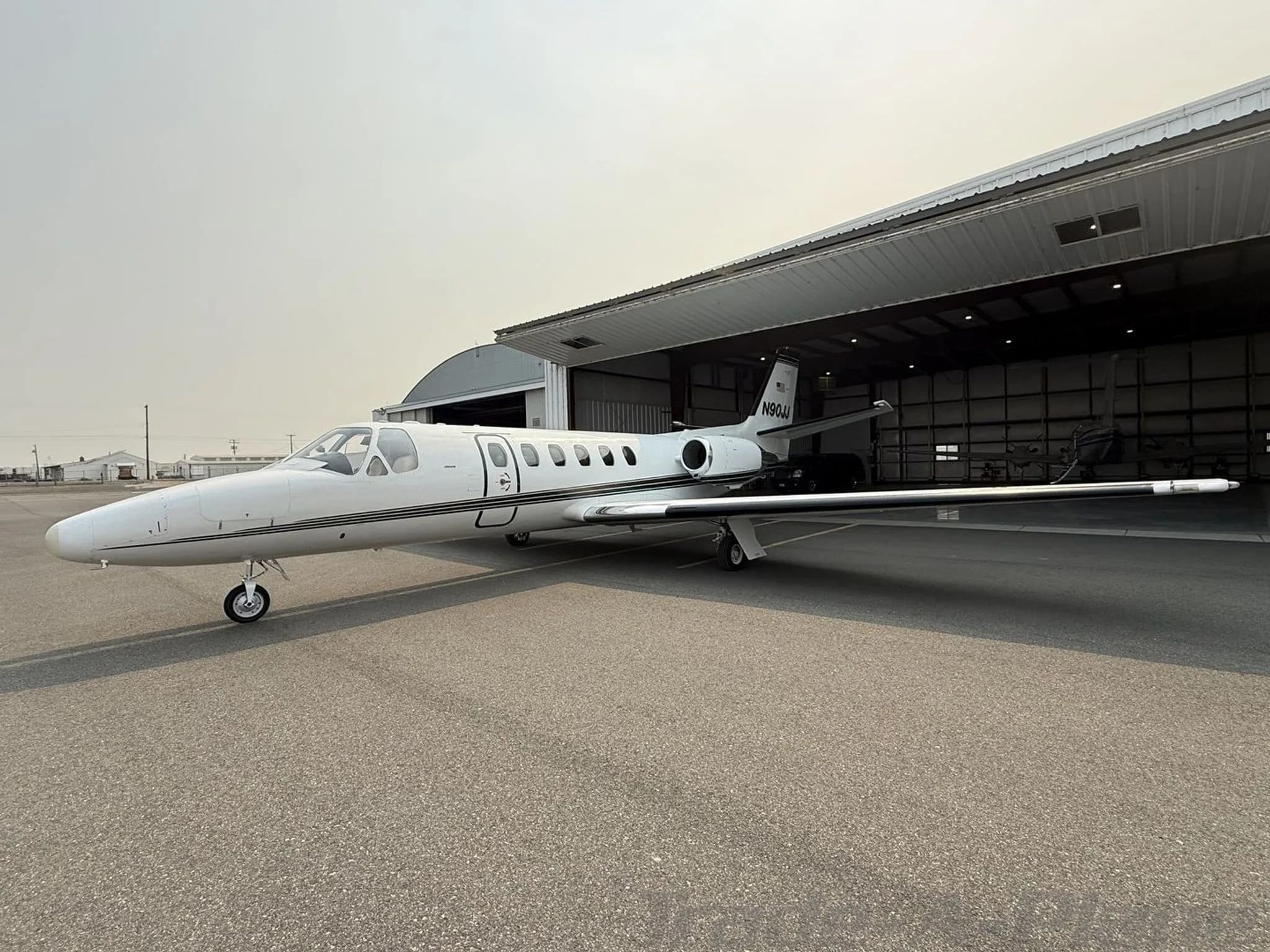 A white Cessna Citation jet, N90JJ, with black and gold stripes, parked on an tarmac next to an open hangar.
