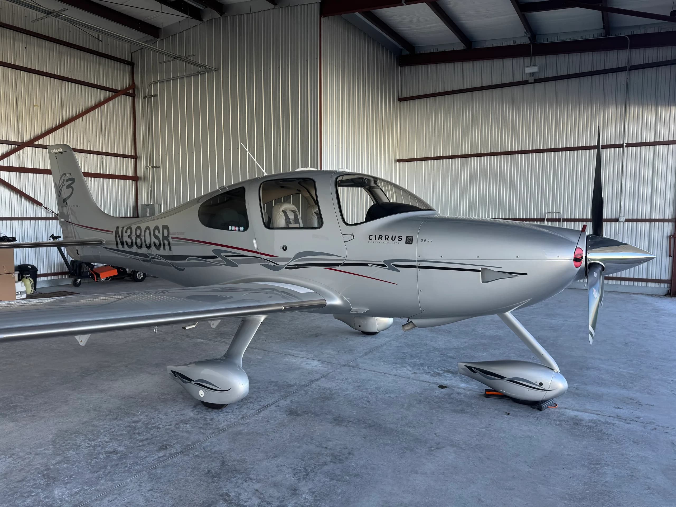 A silver Cirrus SR22 Generation Three aircraft with N380SR registration is parked in a hangar, viewed from the front-right.