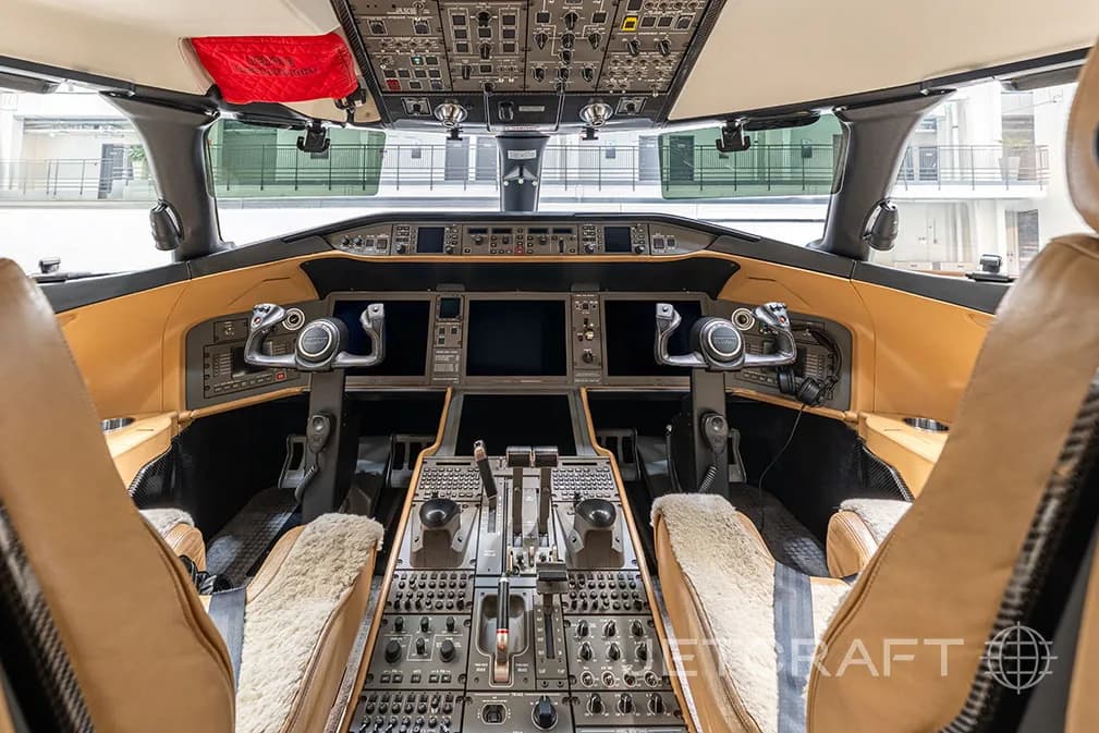 The cockpit of a business jet, featuring multiple display screens, control yokes, and a central throttle quadrant, with tan leather trim.