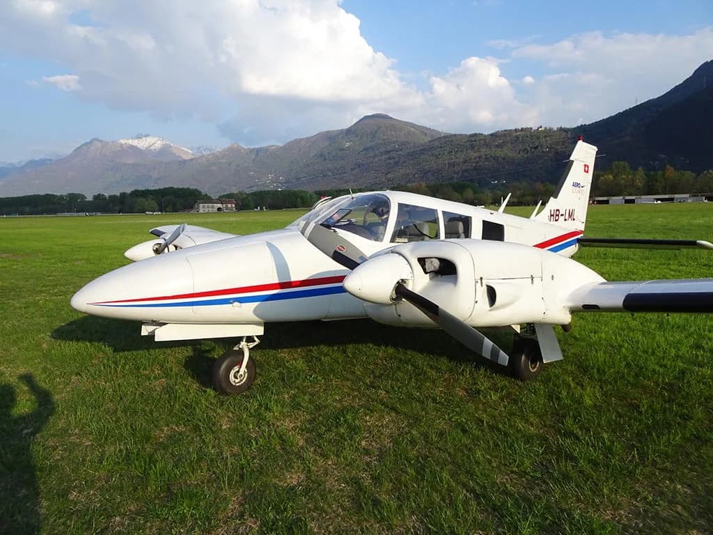 A white twin-engine Piper PA-30 Twin Comanche aircraft with red and blue stripes on a grassy airfield with mountains in the background.
