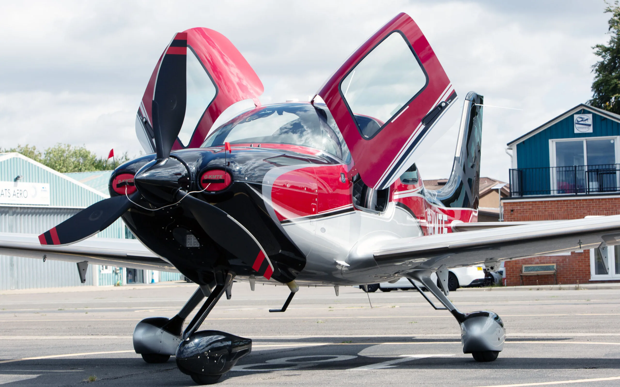 A low-angle front view of a red, black, and silver Cirrus SR22 aircraft with its gull-wing doors open, parked on an airfield.