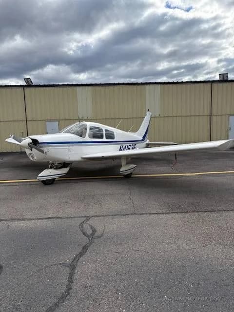 A white single-engine propeller aircraft with blue stripes (N4163F) parked on an asphalt tarmac next to a hangar.