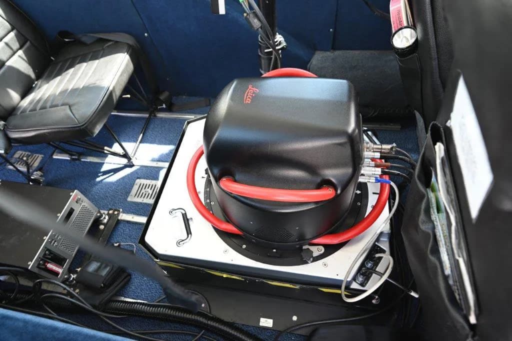 A black Leica sensor unit with red tubing mounted on a platform inside the aircraft cabin, with blue carpet and a seat visible.