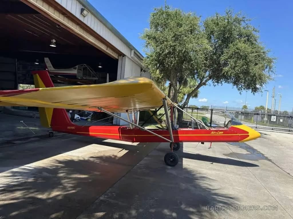 A red and yellow Air-Cam aircraft parked in front of a hangar.