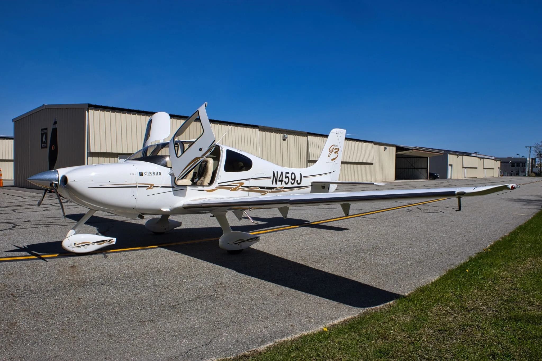 A front-side view of the white Cirrus SR22 GTS Generation Three aircraft (N459J) with both gull-wing doors open, parked near hangars.