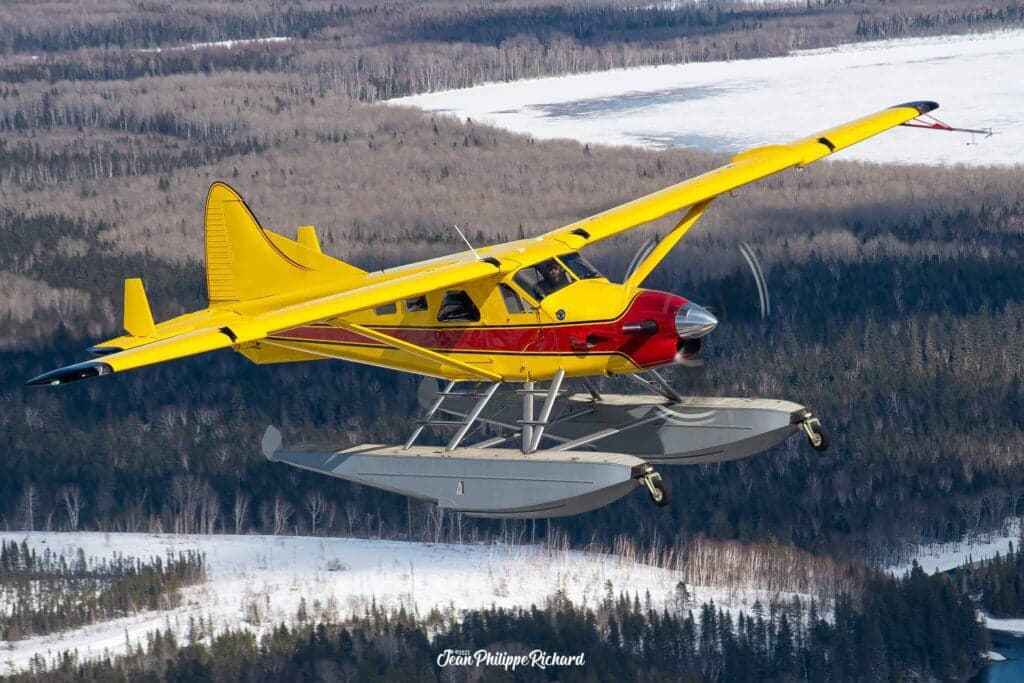 Yellow and red floatplane in flight over a forest and frozen lake.