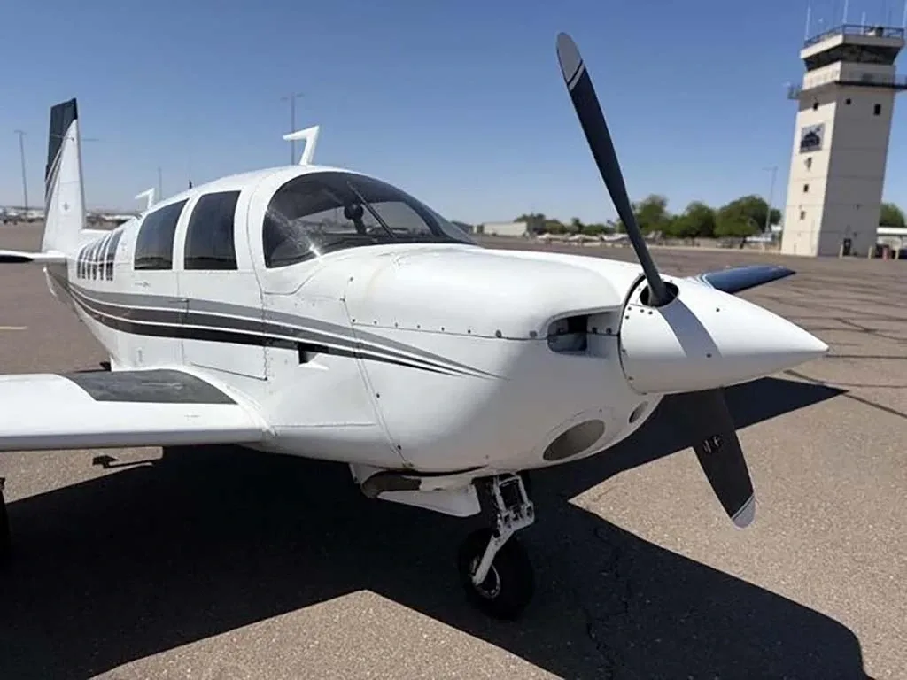 A front-side view of the white Mooney M20 aircraft N2634W parked on an airfield, with an air traffic control tower in the background.