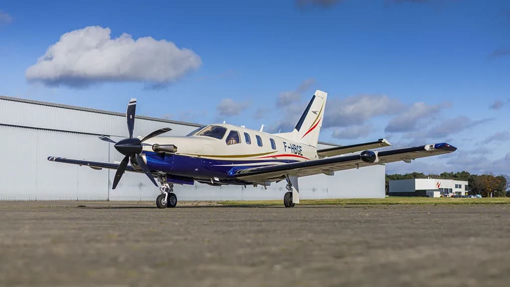 A white and blue TBM aircraft with registration F-HBGE parked on an airfield in front of a hangar.