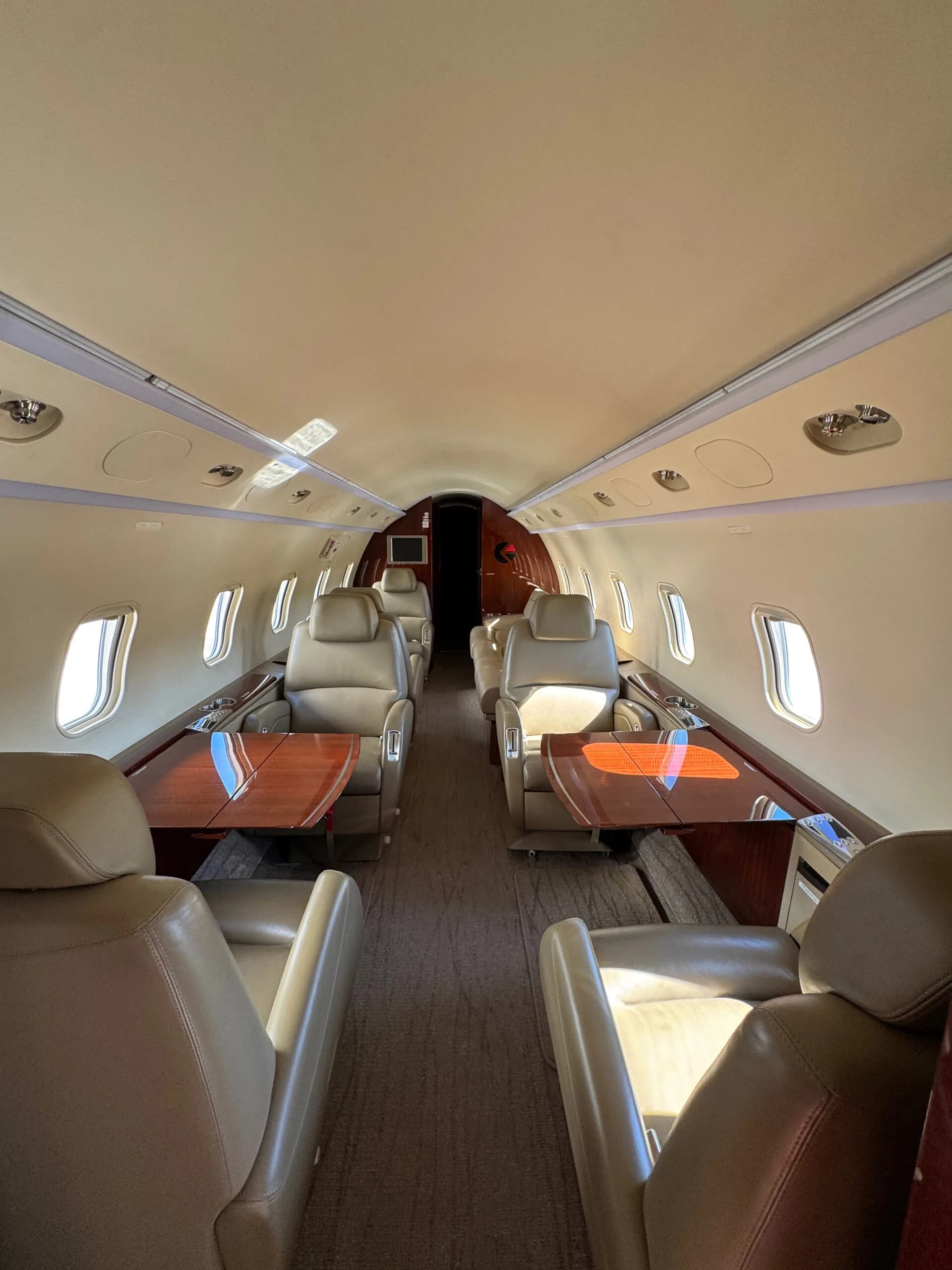 Interior view of the aircraft cabin looking forward, featuring beige leather seats and two polished wooden tables.