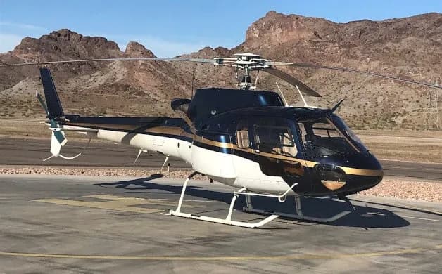 A dark blue and white helicopter with gold stripes is parked on an asphalt pad with mountains in the background.
