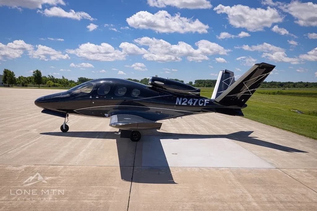 A black Cirrus Vision Jet (N247CF) with a sleek design and silver accents parked on a tarmac with green fields in the background.