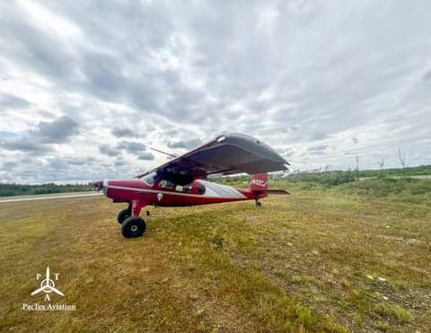 A red and silver single-engine aircraft with large tires, registration N10CJ, parked on a grassy field under a cloudy sky.