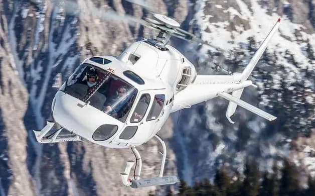 A white helicopter flies in front of a snowy mountain range.
