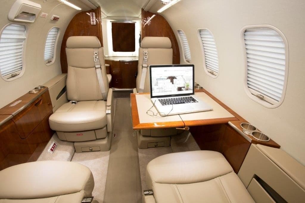 A private jet cabin featuring beige leather seats, wood paneling, and a laptop on a pull-out table.